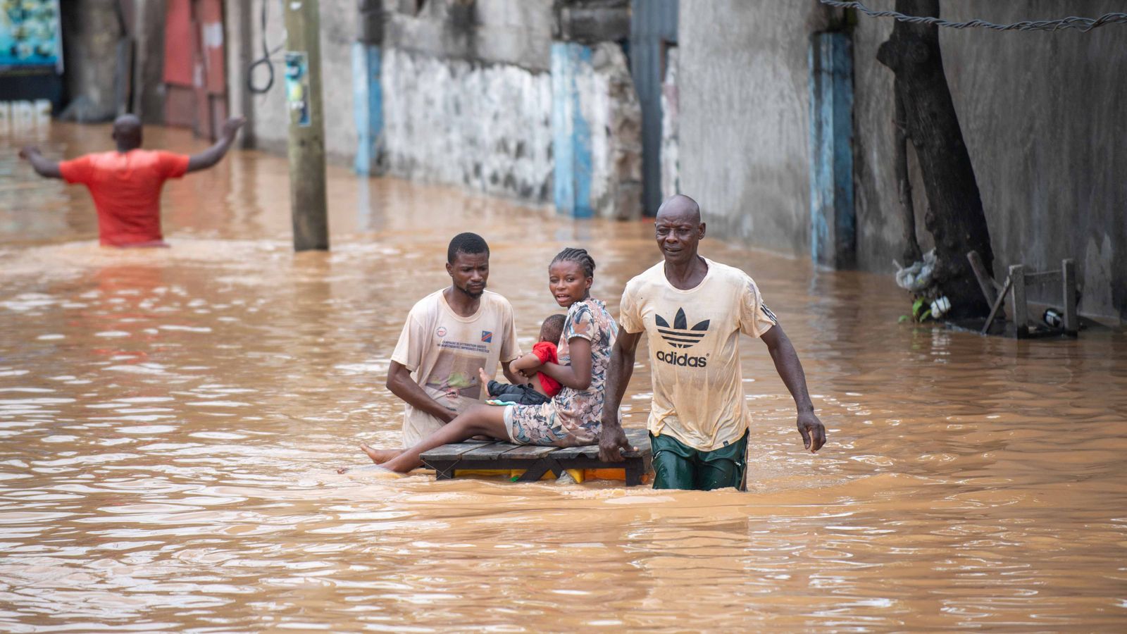 Two men wade through a flooded street pulling a wooden pallet that carries a young woman and a small child