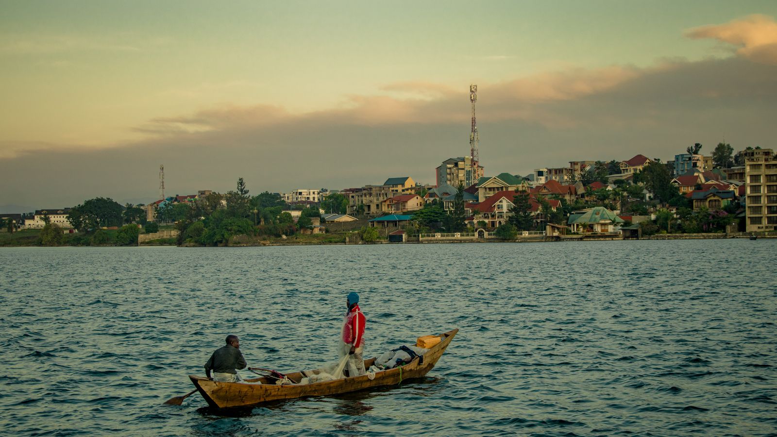 Two fishermen in a wooden canoe on Lake Kivu at dusk, with the town of Goma on the shore behind them