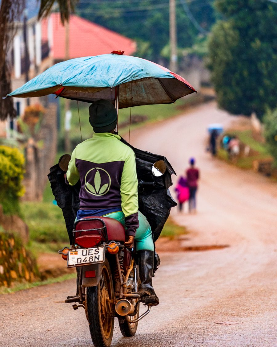 A motorcycle rider holding an umbrella over himself on a wet red-earth road in Uganda